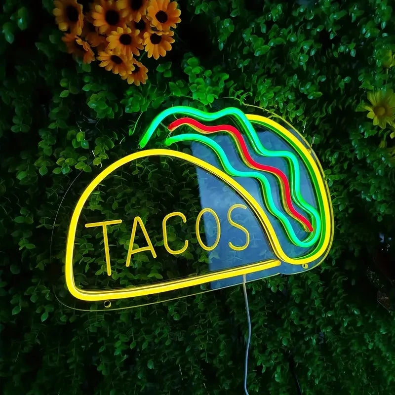 Neon sign shaped like a taco with 'TACOS' text on a green leafy background