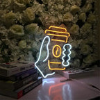 Neon sign of a coffee cup with a straw on a stack of books against a floral background