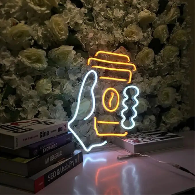 Neon sign of a coffee cup with a straw on a stack of books against a floral background