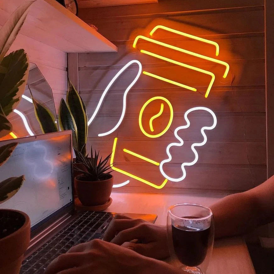 Neon sign of a coffee cup and beans on a wooden wall with a laptop and glass of coffee.