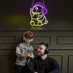 Man and child sitting together with a neon sign of a duck wearing headphones on a dark wall.