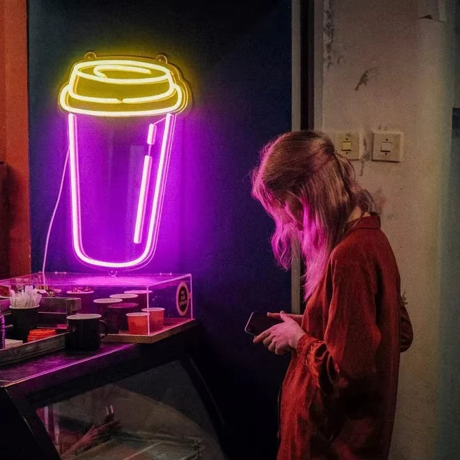 Person standing in front of a neon sign shaped like a coffee cup in a dimly lit room.