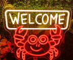Neon sign with a crab design and 'WELCOME' text on a green leafy background.