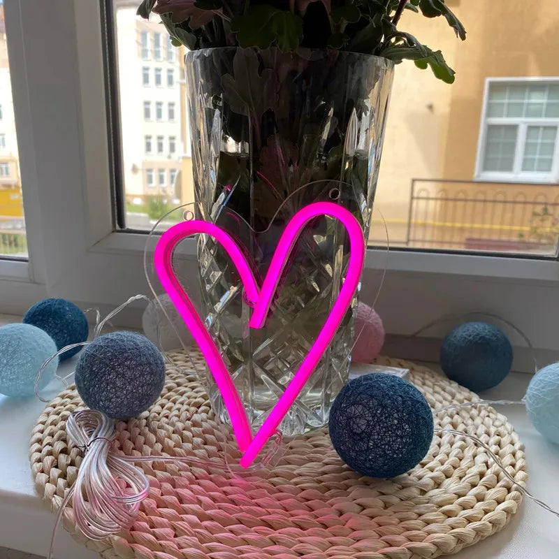 Neon pink heart-shaped light on a woven mat with a vase of flowers and decorative balls.